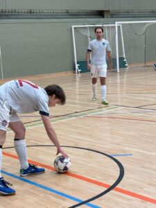 Indoor futsal scene: a player in white bends to touch the ball on a gym floor, another player near the goal in the background.