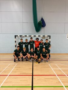 Indoor futsal team photo: 11 players in green jerseys pose with goalie in orange in front of a goal. Team name GRILLA on shirts.