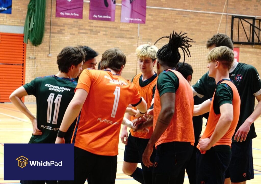 Group of young male athletes in green and orange jerseys huddling in a gym during a team talk; WhichPad logo at bottom-left.