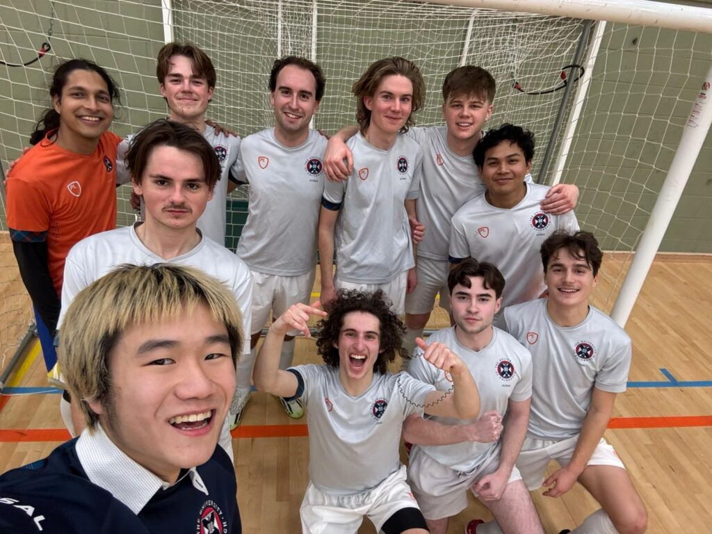 Group of young men in white uniforms posing for a team selfie in an indoor soccer gym, with a goal net behind them.